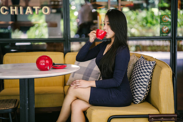 Portrait of a young, attractive, elegant and beautiful Asian woman sitting outdoors in an alfresco cafe and enjoying a hot cup of tea from a red tea set.  - Foto, imagen