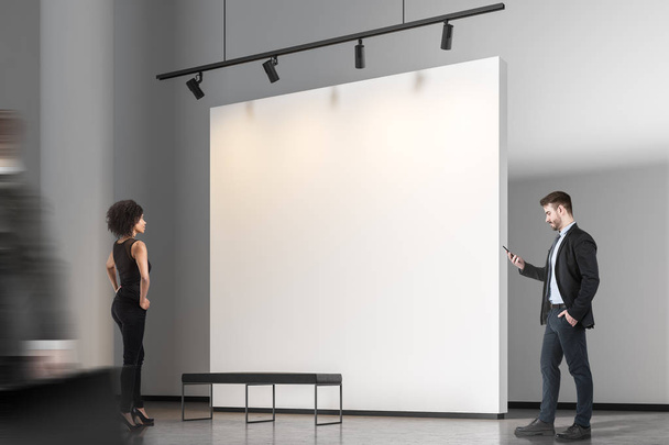 African American woman and bearded businessman with smartphone looking at blank gallery wall. Mock up blurred - Photo, Image