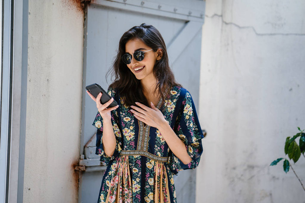 Portrait of a young, attractive and beautiful Indian Asian woman wearing a dress and sunglasses with her smartphone - Photo, image