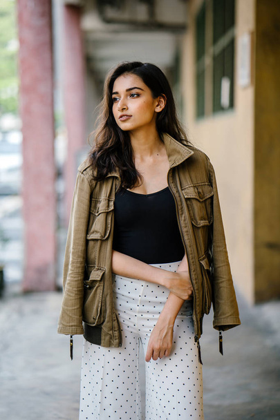 Portrait of a tall, beautiful, slim and elegant Indian Asian woman walking down a walkway in a city in Asia during the day. She is elegantly dressed and has a jacket draped over her shoulders. - Zdjęcie, obraz