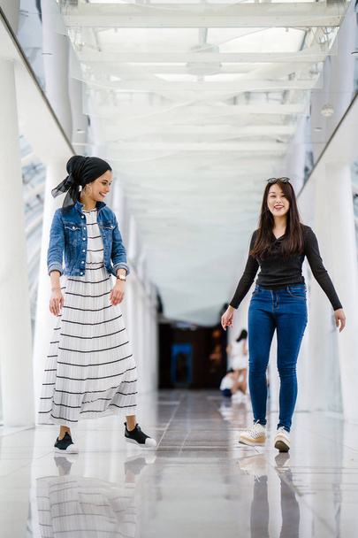 Two friends of different ethnicity stand next to one another on a bridge during the day. One is a Malay Muslim woman and the other is Chinese Asian. They are both young, energetic and attractive. - Фото, изображение