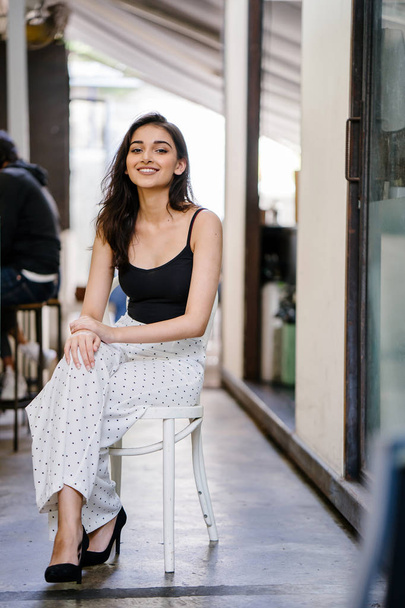 Portrait of a tall, elegant and beautiful Indian Asian girl in a black top and long white pants sitting on a chair in a cafe in the city during the day - Zdjęcie, obraz