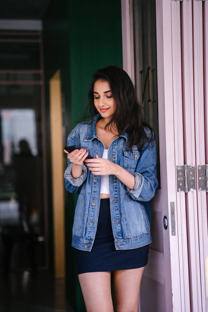 Portrait of a beautiful, tall, elegant and young Indian Asian woman texting on her smartphone. She is wearing a retro denim jacket  - Foto, Imagen