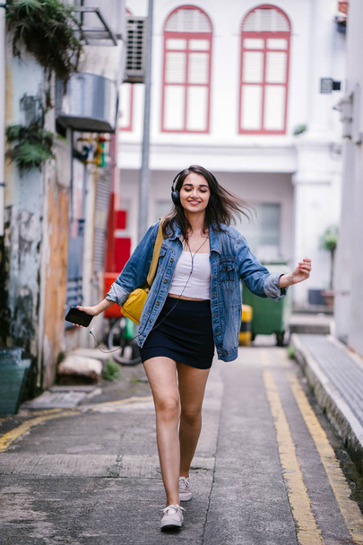 Portrait of a young and attractive Indian Asian woman in a denim jacket and yellow backpack enjoying music on her headphones - Foto, Imagen