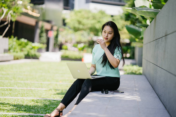 A young and attractive Asian woman student sits and works on her laptop computer on a university campus during the day. She is casually dressed as she studies.  - Zdjęcie, obraz