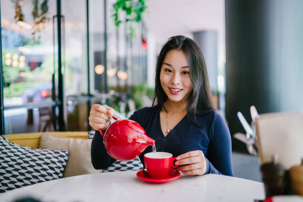 Portrait of a young, attractive, elegant and beautiful Asian woman sitting outdoors in an alfresco cafe and enjoying a hot cup of tea from a red tea set.  - Foto, Imagen