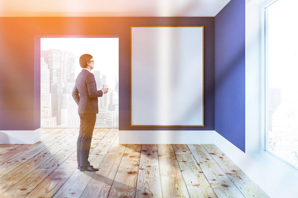 Interior of empty room with blue walls, wooden floor, large windows with a cityscape. Man looking at big vertical poster on the wall. Toned image mock up - Photo, Image
