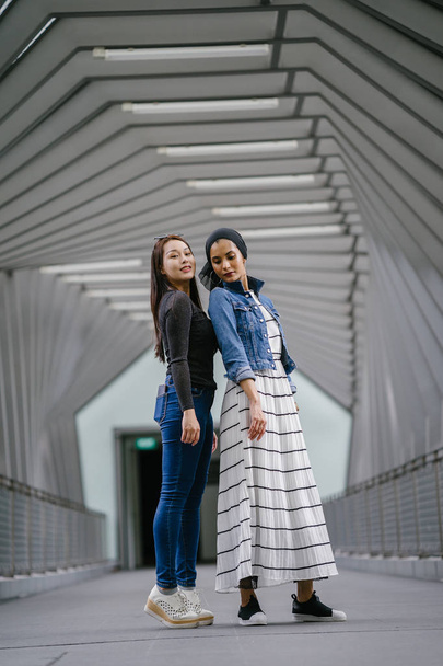 Two friends of different ethnicity on a bridge during the day. One is a Malay Muslim woman and the other is Chinese Asian. They are both young, energetic and attractive. - Foto, immagini