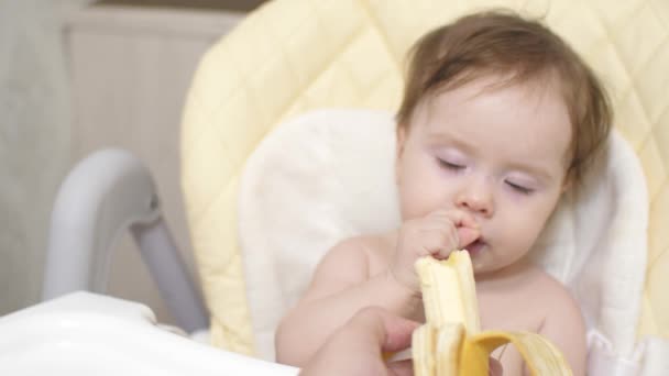 Μικρό μωρό τρώει μια μπανάνα που κάθεται στην καρέκλα στην κουζίνα. - Πλάνα, βίντεο