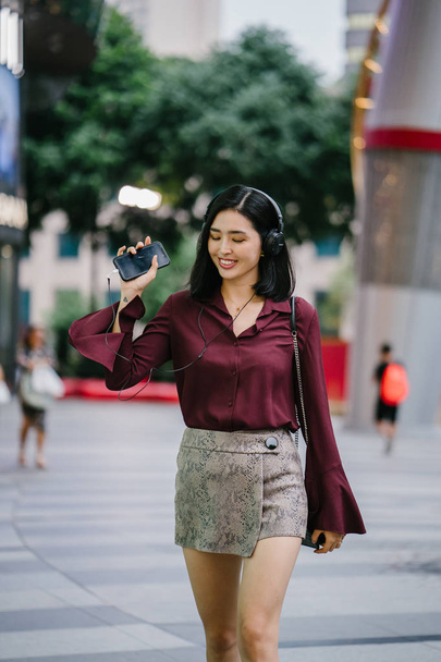 beautiful Korean woman in an elegant blouse and shorts dancing as she walks down a street in the city with her headphones on - Photo, image
