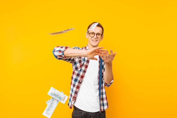 Portrait of a happy man, wearing glasses and a plaid shirt, throwing out money banknotes on a yellow background - 写真・画像