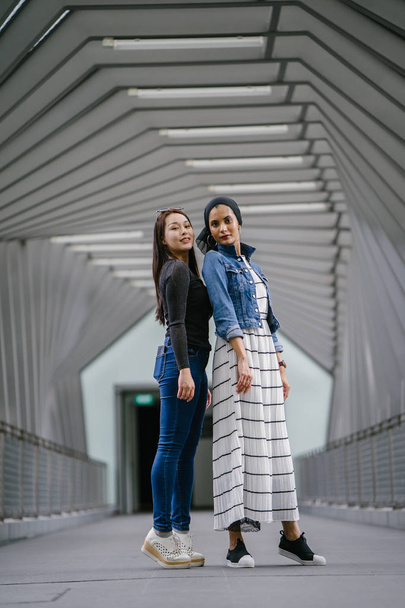 Two friends of different ethnicity on a bridge during the day. One is a Malay Muslim woman and the other is Chinese Asian. They are both young, energetic and attractive. - Zdjęcie, obraz