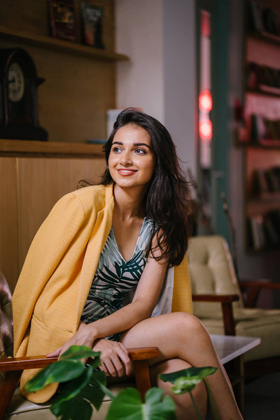  fashionable, beautiful and young Indian Asian woman in a dress and a yellow jacket draped over her shoulders. She is sitting on a chair in a stylish interior - Foto, Bild