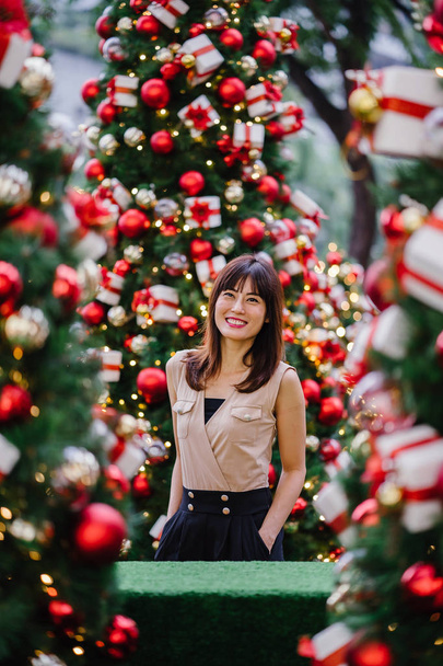 Chinese Asian tourist woman smiles and poses for a portrait with beautifully decorated Christmas trees during the day in the city - Fotografie, Obrázek