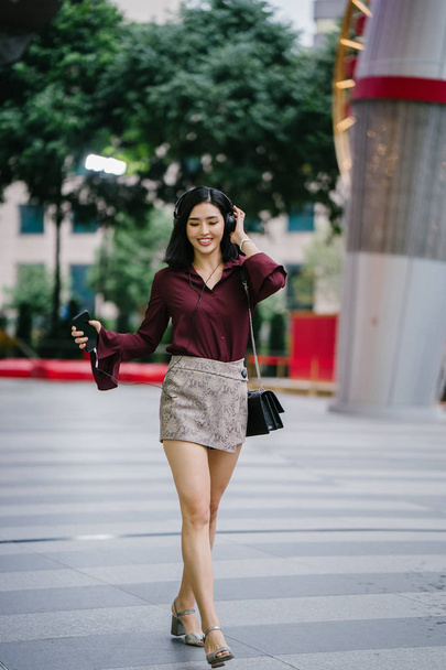 beautiful Korean woman in an elegant blouse and shorts dancing as she walks down a street in the city with her headphones on - 写真・画像
