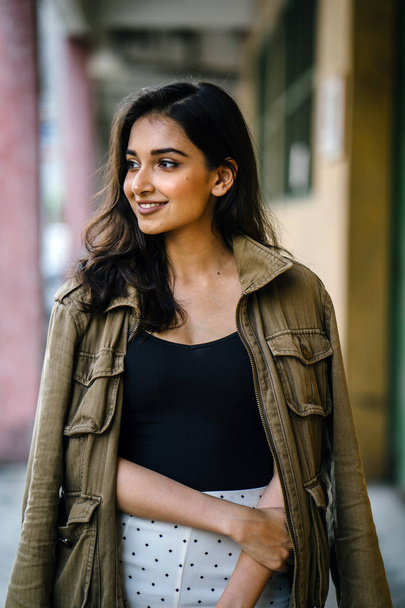 Portrait of a tall, beautiful, slim and elegant Indian Asian woman walking down a walkway in a city in Asia during the day. She is elegantly dressed and has a jacket draped over her shoulders. - Foto, Imagem