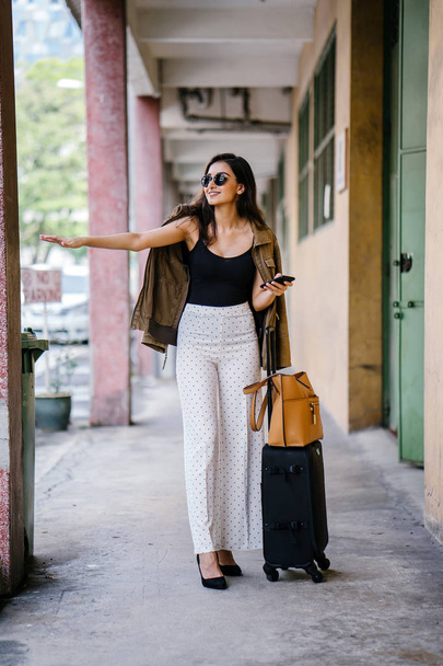 young and attractive Asian Indian woman books a ride through her ride hailing app on her smartphone. She is standing in a walkway with her luggage and is stylishly dressed in work wear and shades - Zdjęcie, obraz