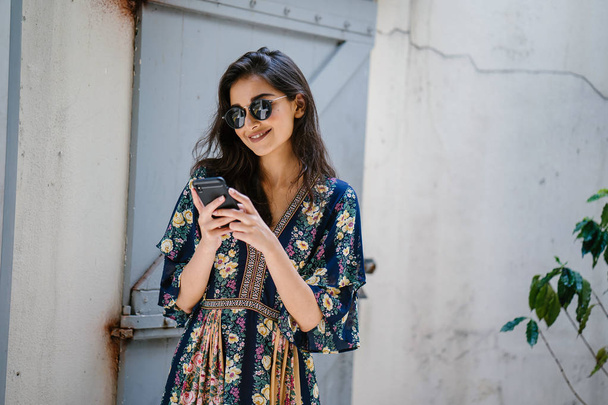 Portrait of a young, attractive and beautiful Indian Asian woman wearing a dress and sunglasses with her smartphone - Foto, Imagem