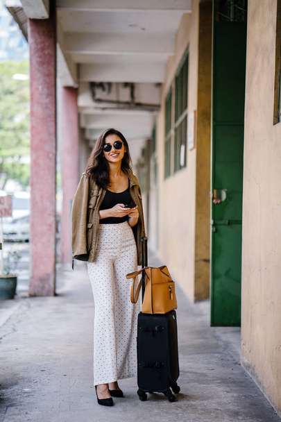 young and attractive Asian Indian woman books a ride through her ride hailing app on her smartphone. She is standing in a walkway with her luggage and is stylishly dressed in work wear and shades - 写真・画像