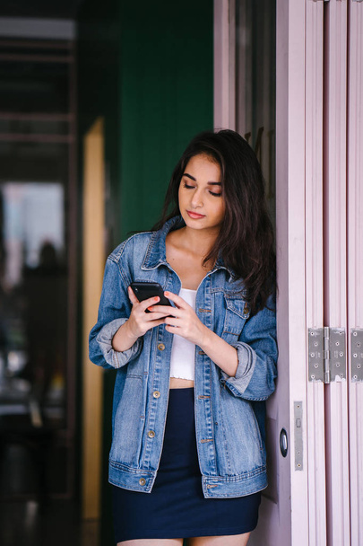 Portrait of a beautiful, tall, elegant and young Indian Asian woman texting on her smartphone. She is wearing a retro denim jacket  - Foto, Imagem