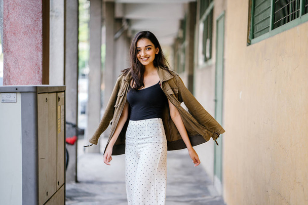 Portrait of a tall, beautiful, slim and elegant Indian Asian woman walking down a walkway in a city in Asia during the day. She is elegantly dressed and has a jacket draped over her shoulders. - Zdjęcie, obraz