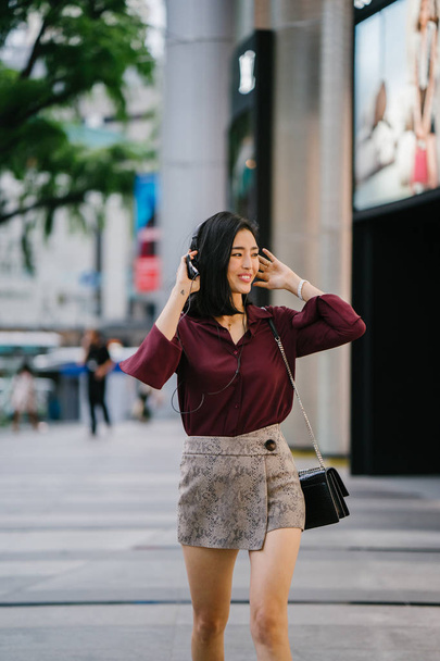 beautiful Korean woman in an elegant blouse and shorts dancing as she walks down a street in the city with her headphones on - Fotografie, Obrázek