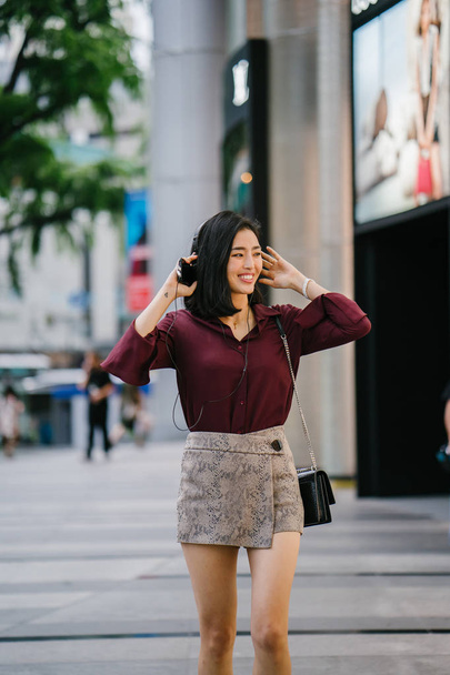 beautiful Korean woman in an elegant blouse and shorts dancing as she walks down a street in the city with her headphones on - Foto, Bild