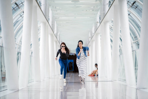 Two diverse friends sprint down a well-lit white futuristic overpass bridge towards the camera. One is a Muslim Middle Eastern woman and the other is Chinese. - Photo, image