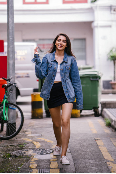 tall, young and elegant Indian Asian woman walking down an alley in the city during the day. She is wearing a retro denim jacket - Фото, зображення