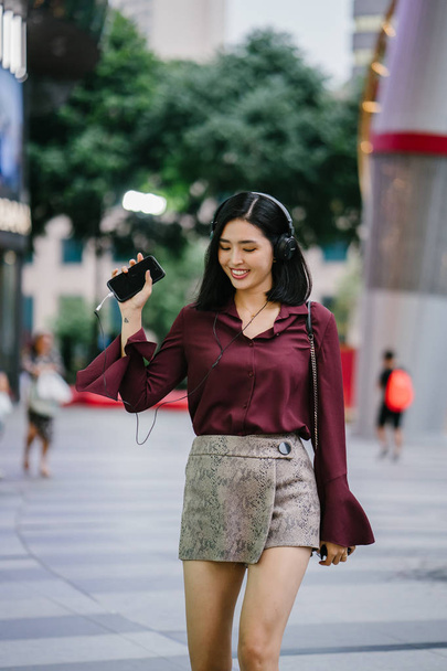 beautiful Korean woman in an elegant blouse and shorts dancing as she walks down a street in the city with her headphones on - Foto, afbeelding