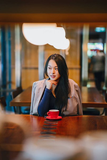 Portrait of a young, attractive, elegant and beautiful Asian woman sitting outdoors in an alfresco cafe and enjoying a hot cup of tea from a red tea set.  - Foto, Imagem