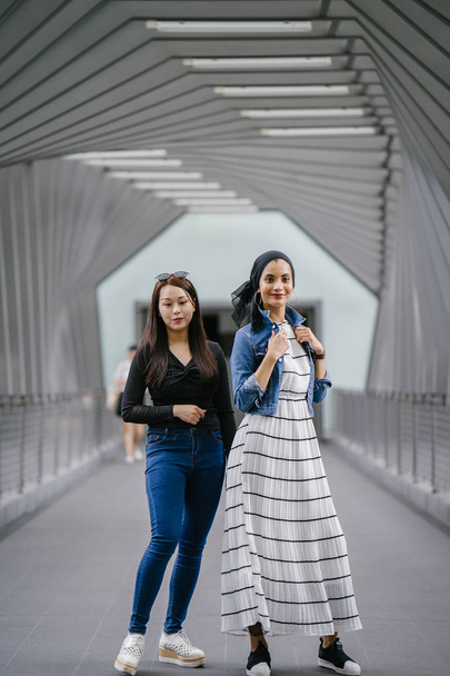 Two friends of different ethnicity on a bridge during the day. One is a Malay Muslim woman and the other is Chinese Asian. They are both young, energetic and attractive. - Фото, зображення