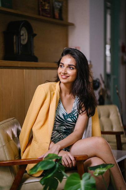  fashionable, beautiful and young Indian Asian woman in a dress and a yellow jacket draped over her shoulders. She is sitting on a chair in a stylish interior - Fotó, kép