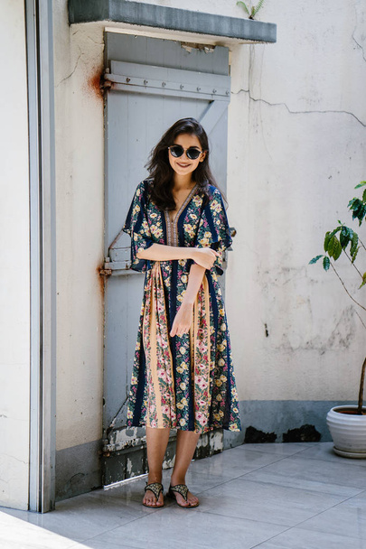 magas, elegáns és gyönyörű indiai ázsiai nő egy nyári ruha és napszemüveg mosolyog mint ő jelent egy utcában, egy sikátorban a nap folyamán - Fotó, kép