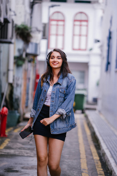Portrait of a young and attractive Indian Asian woman in a denim jacket and yellow backpack enjoying music on her headphones - Foto, Imagen