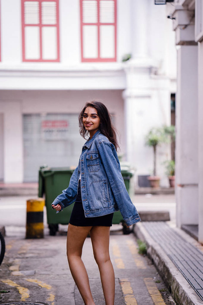 tall, young and elegant Indian Asian woman walking down an alley in the city during the day. She is wearing a retro denim jacket - Foto, afbeelding