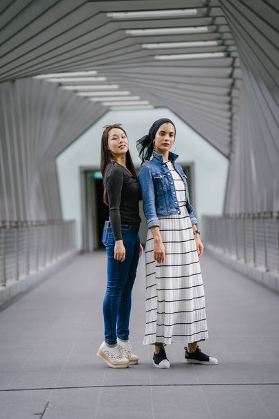 Two friends of different ethnicity on a bridge during the day. One is a Malay Muslim woman and the other is Chinese Asian. They are both young, energetic and attractive. - Foto, afbeelding