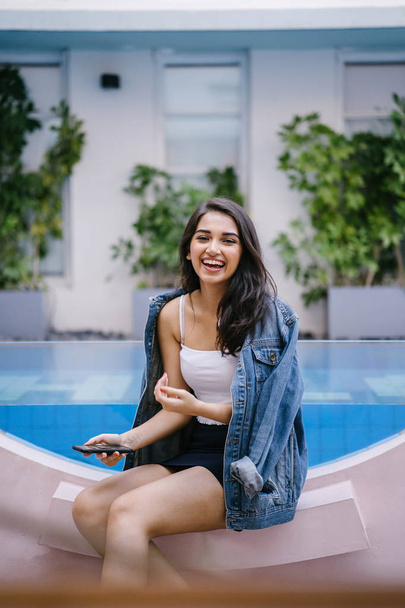 A lifestyle portrait of a young Indian Asian woman sitting by a resort pool during the day while holding smartphone - 写真・画像