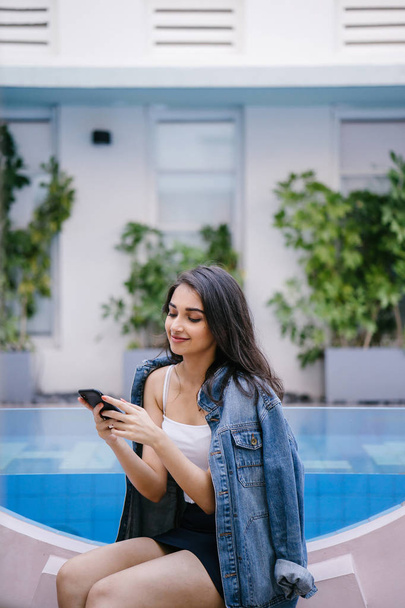 A lifestyle portrait of a young Indian Asian woman sitting by a resort pool during the day while holding smartphone - Valokuva, kuva