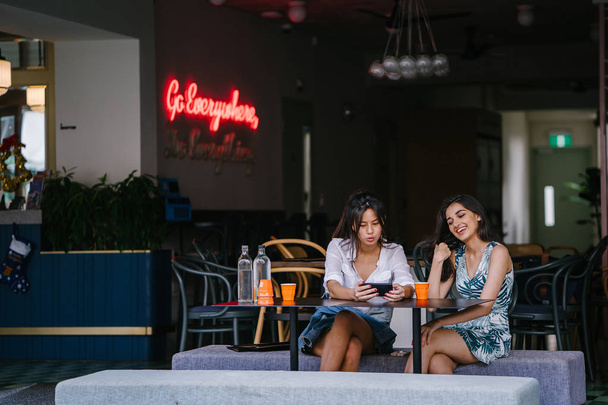 Dwóch bliskich przyjaciół (kobiety) siedzieć w kawiarni w ciągu dnia i Obejrzyj film razem na smartfonie. Są zarówno uśmiechnięte jak cieszyć się swoim towarzystwem i wideo. - Zdjęcie, obraz