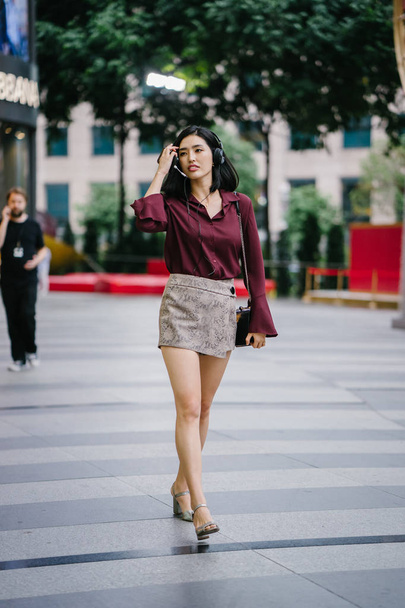 beautiful Korean woman in an elegant blouse and shorts dancing as she walks down a street in the city with her headphones on - 写真・画像