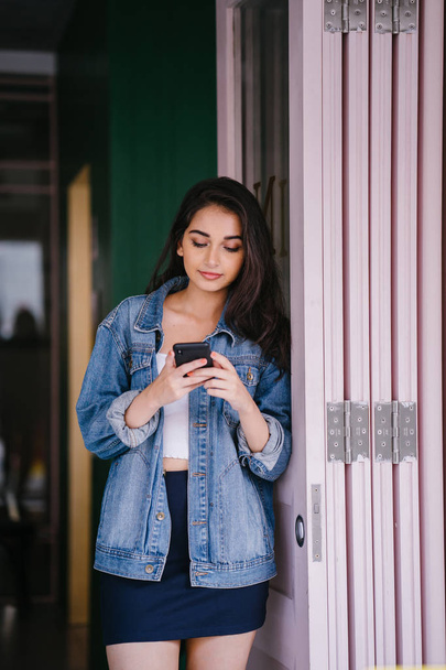 Portrait of a beautiful, tall, elegant and young Indian Asian woman texting on her smartphone. She is wearing a retro denim jacket  - Φωτογραφία, εικόνα