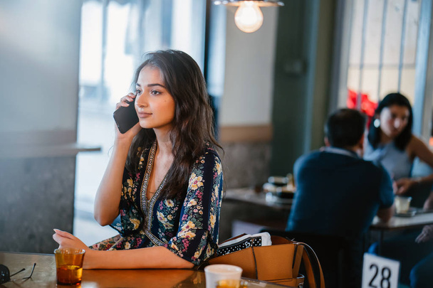 Portrait of a young and attractive Indian Asian woman frowning as she talks on her smartphone in a cafe during the day - 写真・画像