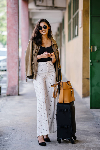 young and attractive Asian Indian woman books a ride through her ride hailing app on her smartphone. She is standing in a walkway with her luggage and is stylishly dressed in work wear and shades - Zdjęcie, obraz