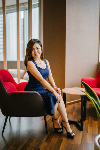 A pretty, young businesswoman sits in a red armchair in her office. She is Asian Chinese and wearing a blue dress and is smiling as she poses for her head shot. - Foto, imagen