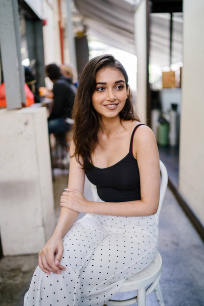 Portrait of a tall, elegant and beautiful Indian Asian girl in a black top and long white pants sitting on a chair in a cafe in the city during the day - Foto, Bild