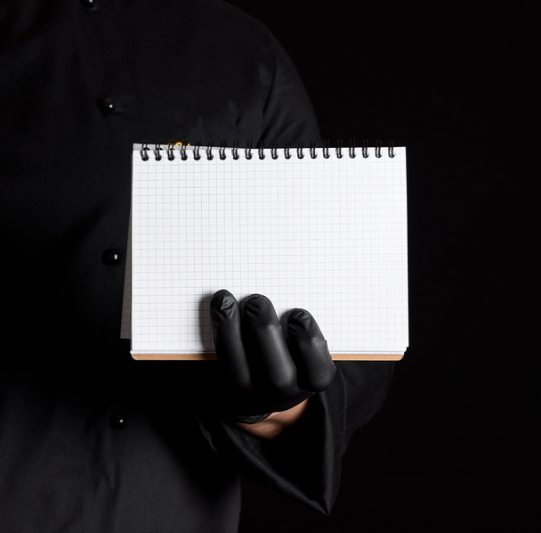 Chef-kok in zwart uniform en zwarte latex handschoenen houden een lege notebook, zwarte achtergrond - Foto, afbeelding