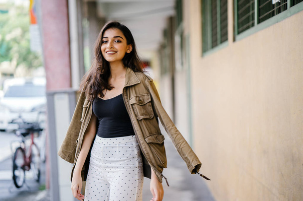 Portrait of a tall, beautiful, slim and elegant Indian Asian woman walking down a walkway in a city in Asia during the day. She is elegantly dressed and has a jacket draped over her shoulders. - Foto, immagini