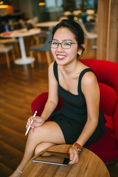 昼間同僚のオフィスに座っているときに、スタイラスペンでタブレットデバイスを操作している若い美しいアジア人女性のビジネスポートレート. - 写真・画像