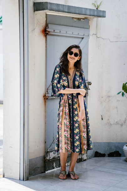 alta, elegante, e bella donna asiatica indiana in un abito estivo e occhiali da sole sorride mentre posa su una strada in un vicolo durante il giorno
 - Foto, immagini